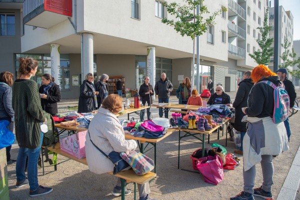 Auf dem Bild sieht man einen großen Tisch mit einem Kleidertausch. Herum stehen einige Menschen, die Kaffee und Tee trinken